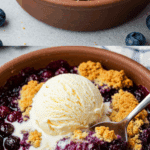 A close-up of a warm bowl of Easy Blueberry Cobbler with a scoop of vanilla ice cream melting on top.