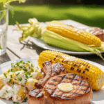 A plated meal of a grilled pork chop with potato salad and corn on the cob.