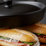 Two wrapped Pan Bagnat sandwiches being pressed under a heavy cast-iron skillet.