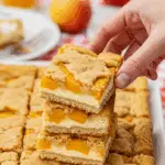 A hand taking a delicious Peach Cobbler Cheesecake Bar from a platter at a summer barbecue.