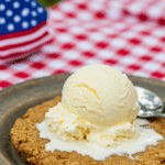 A perfect slice of warm homemade Peach Pie with a large scoop of melting vanilla ice cream at a summer picnic.