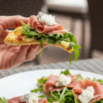 A hand holding a slice of the finished Prosciutto Arugula Flatbread.