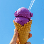 A hand holding a delicious ice cream cone with a large scoop of vibrant purple ube ice cream.