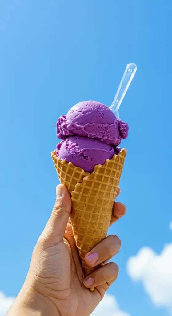 A hand holding a delicious ice cream cone with a large scoop of vibrant purple ube ice cream.