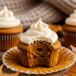 A hand holding a delicious, homemade Pumpkin Cupcake with a bite taken out, in a cozy fall setting.