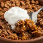 A close-up of a warm bowl of Pecan Pumpkin Crunch with a large dollop of bourbon whipped cream melting on top.