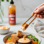 A hand using chopsticks to dip a piece of homemade, crispy sesame tofu into a small bowl of creamy sriracha mayo.