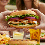 A hand holding a large, delicious, and fully assembled homemade Chicken Bacon Swiss Sandwich at a summer barbecue.