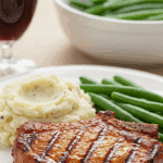 A classic dinner plate with a thick, saucy BBQ pork chop served with mashed potatoes and green beans.